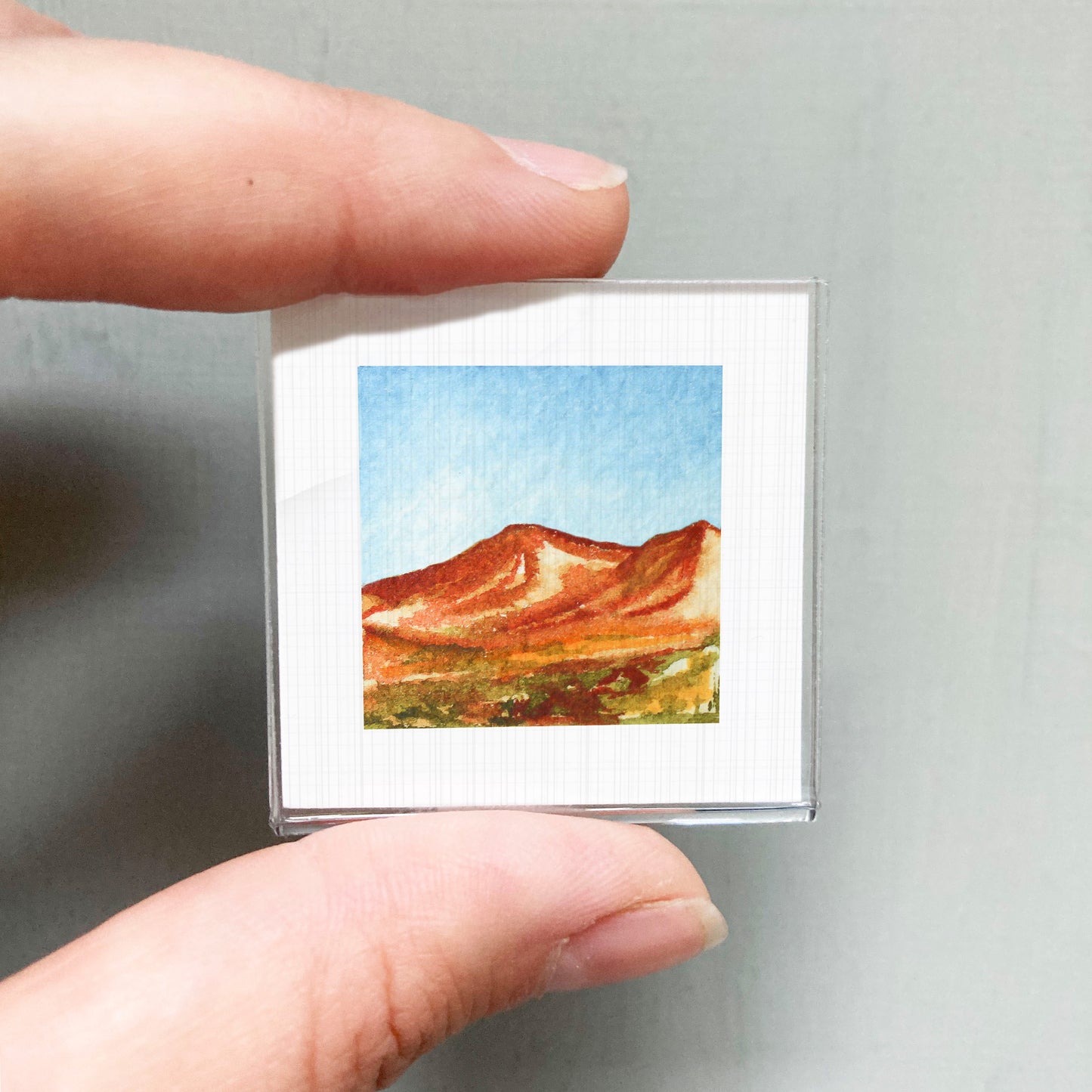 A person's hand holding a small square picture of a mountain landscape.