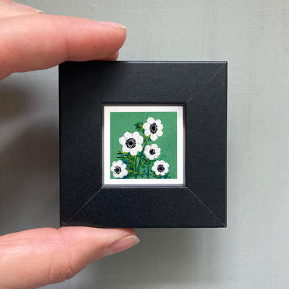 A person's hand holding a small square picture frame containing a painting of white flowers against a green background.