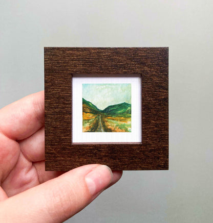 A hand is holding a small, square-shaped wooden frame containing a miniature painting of a landscape with mountains and a winding road.