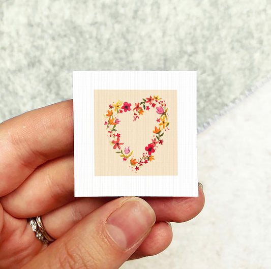 A hand holding a small square card with a heart-shaped floral design on it.