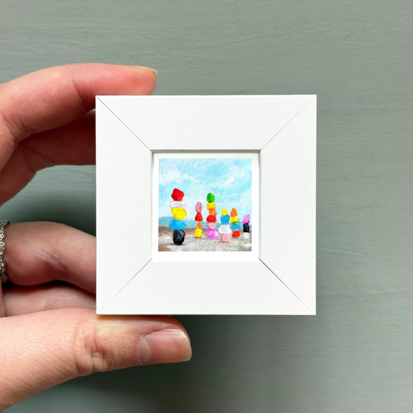 A hand is holding a small white frame containing a colorful painting of a beach scene with people and various objects.