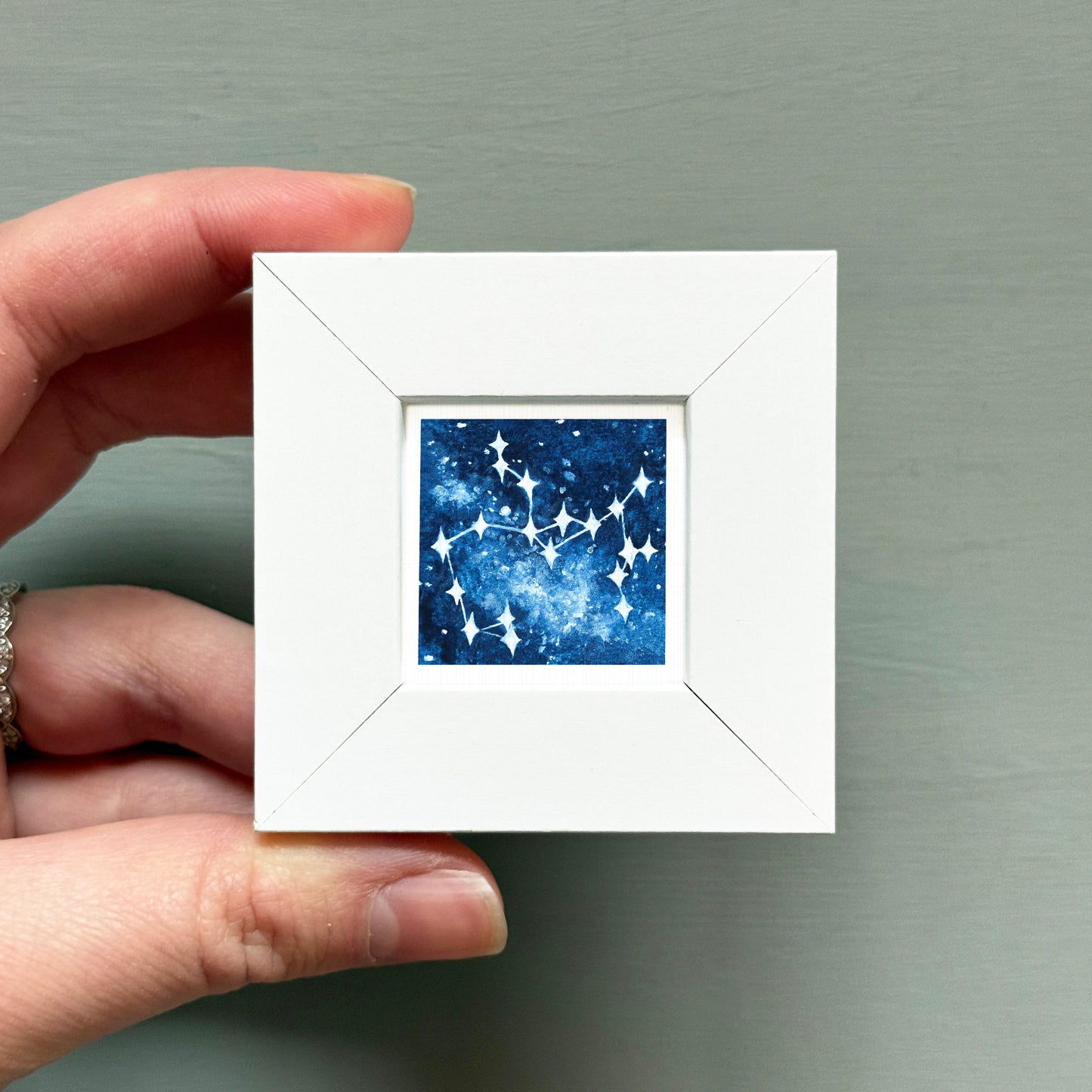 A hand is holding a small white square frame containing a painting of a starry night sky.