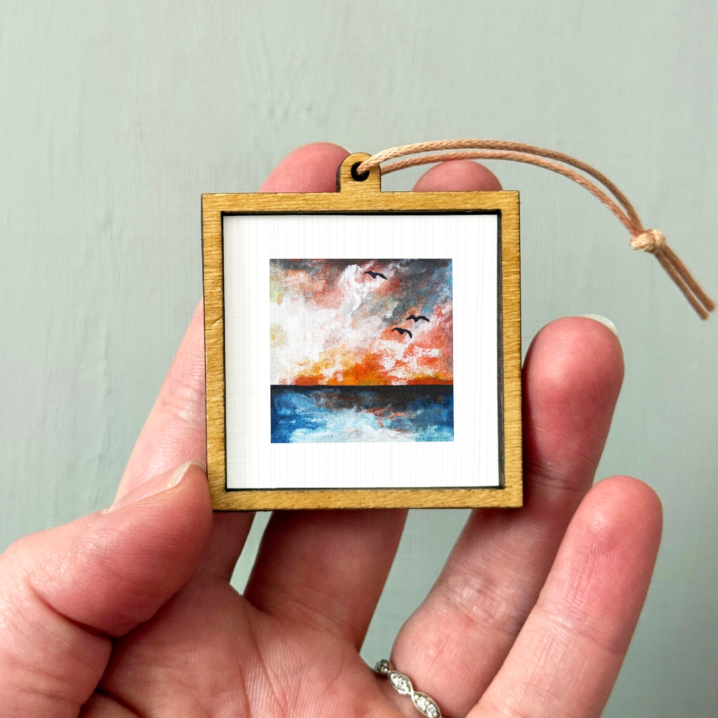 A hand holding a small wooden frame containing a painting of a sunset over a body of water.