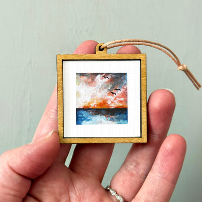 A hand holding a small wooden frame containing a painting of a sunset over a body of water.