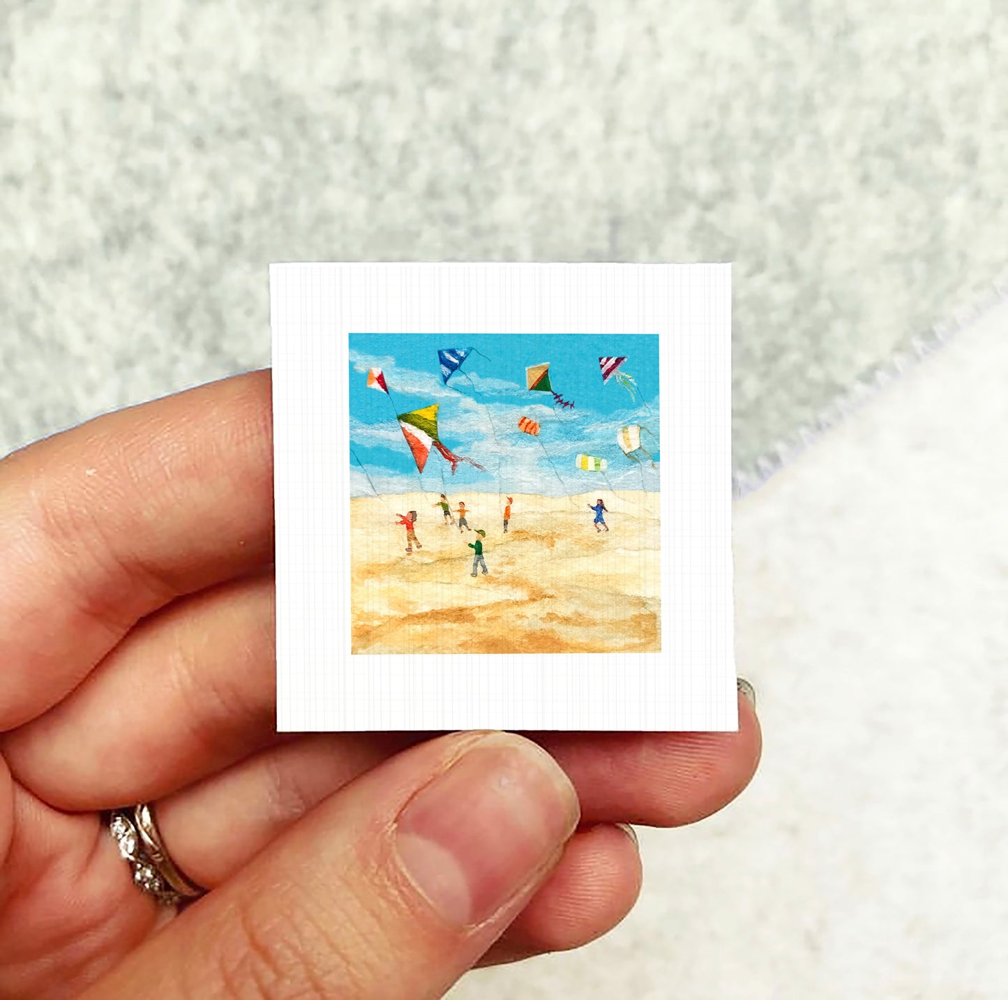 A hand holding a small square picture depicting a beach scene with people flying colorful kites.
