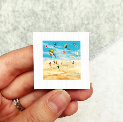 A hand holding a small square picture depicting a beach scene with people flying colorful kites.