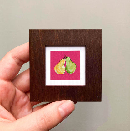 A hand is holding a small square frame containing a painting of two pears.