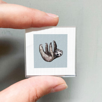 A person's hand is holding a small square picture of a sloth.