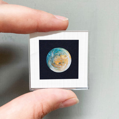 A person's hand is holding a small square frame containing a detailed image of a planet or celestial body.