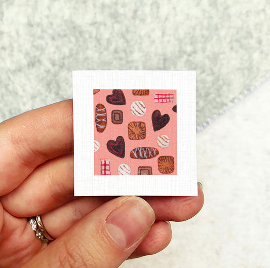 A hand holding a small square with a pattern of hearts and other shapes on a pink background.