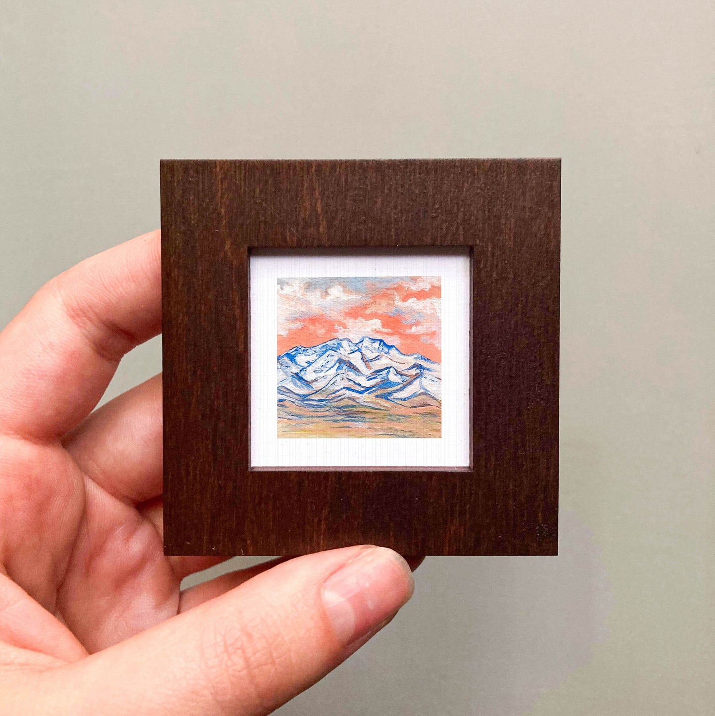 A hand is holding a small wooden frame containing a miniature painting of a mountain landscape.