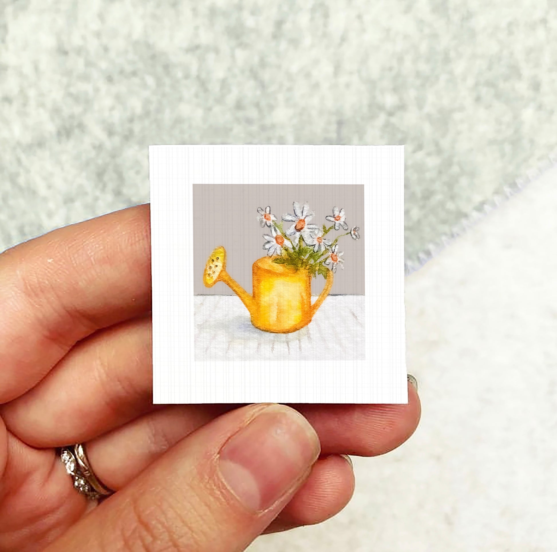 A hand holding a small square card with a painting of a yellow watering can filled with white flowers.