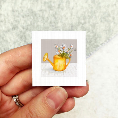 A hand holding a small square card with a painting of a yellow watering can filled with white flowers.