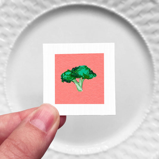 A hand is holding a small square with a picture of a green broccoli on a red background.