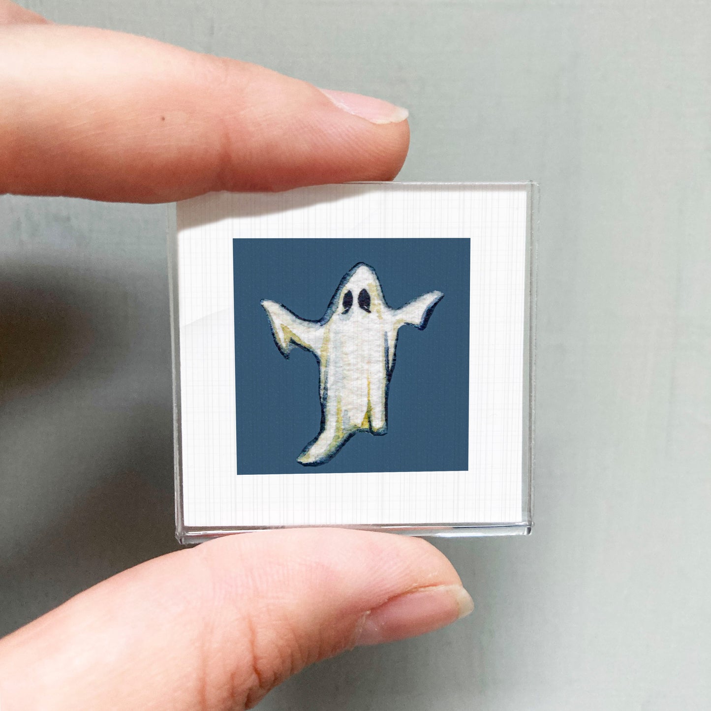 A person's hand is holding a small square picture of a ghost against a blue background.