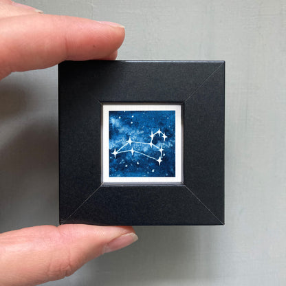 A hand is holding a small black square frame containing a framed image of a starry night sky with a constellation.