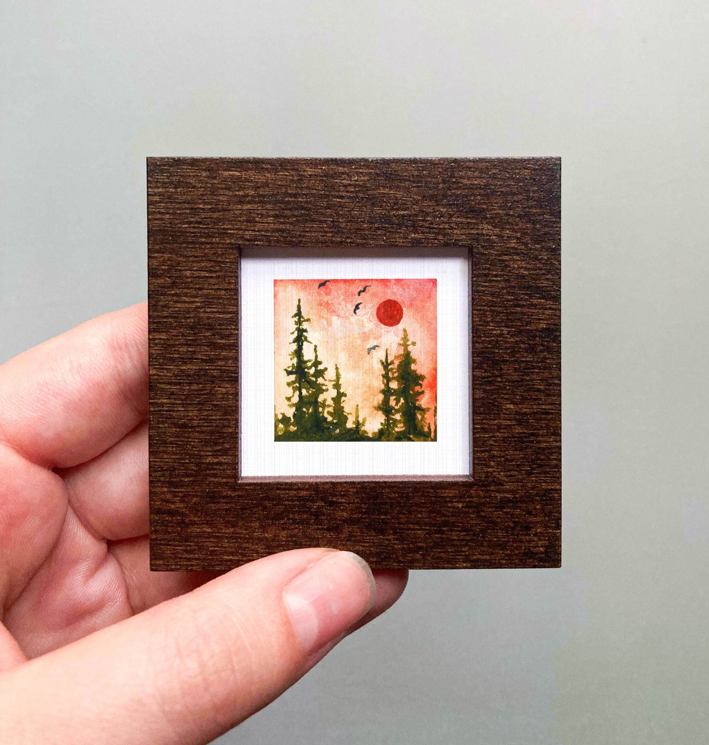 A hand is holding a small framed picture of a forest scene with a red sun in the sky.