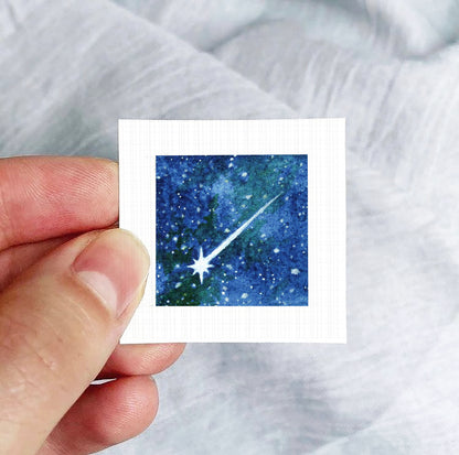 A hand is holding a small square-shaped paper with a watercolor painting of a starry night sky on it.