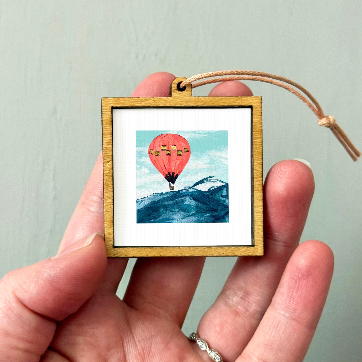 A person's hand holding a small wooden frame containing a framed image of a hot air balloon over a mountain landscape.