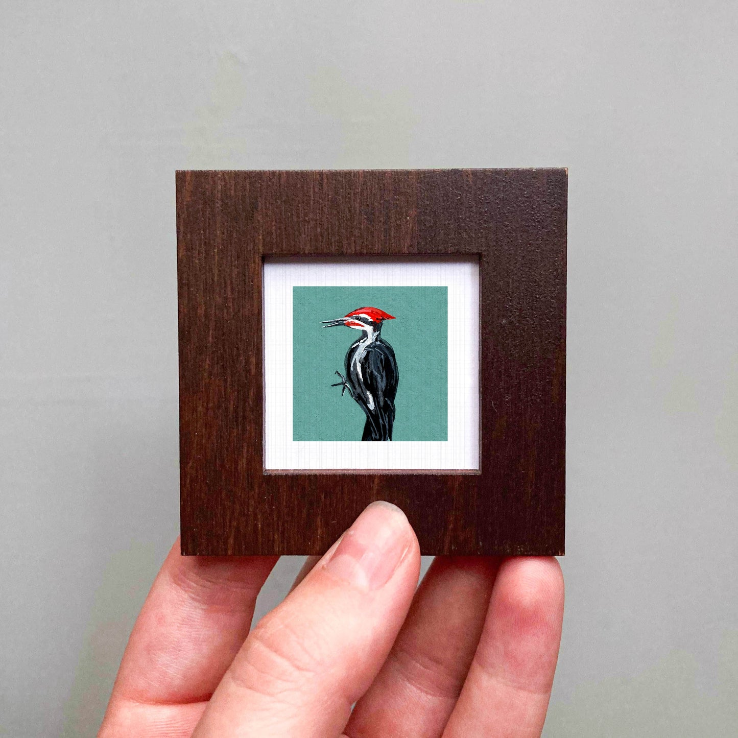 A person's hand is holding a small wooden frame containing a framed image of a woodpecker.