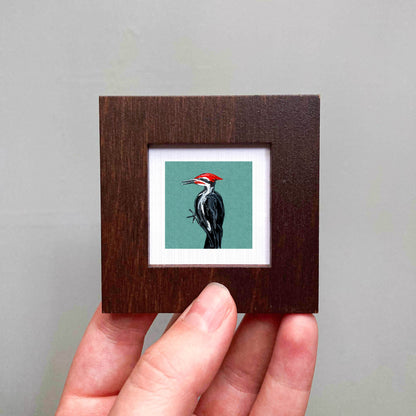 A person's hand is holding a small wooden frame containing a framed image of a woodpecker.