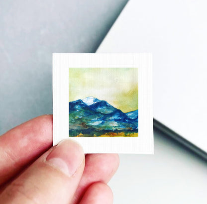 A hand is holding a small square artwork depicting a mountain landscape with a blue sky and green mountains.