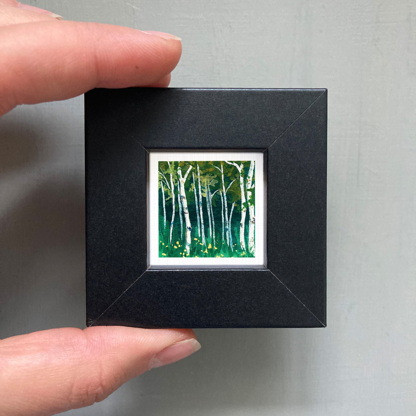 A person's hand holding a small, square-shaped black frame containing a miniature painting of a forest scene with white birch trees and green foliage.