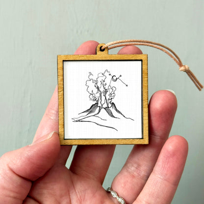 A hand holding a small wooden frame containing a black and white line drawing of a mountain landscape.