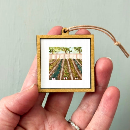 A hand holding a small wooden frame containing a painting of a garden with rows of plants.