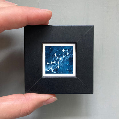 A hand is holding a small black frame containing a framed image of a starry night sky.