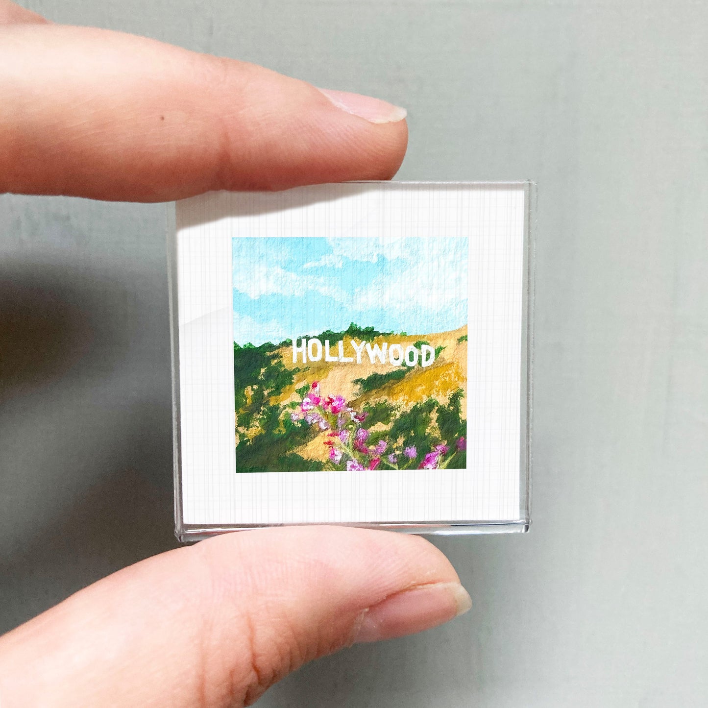A person's hand holding a small square picture of a scenic landscape with the word "Hollywood" written on it.
