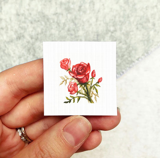 A hand holding a small square card with a painting of red roses on it.