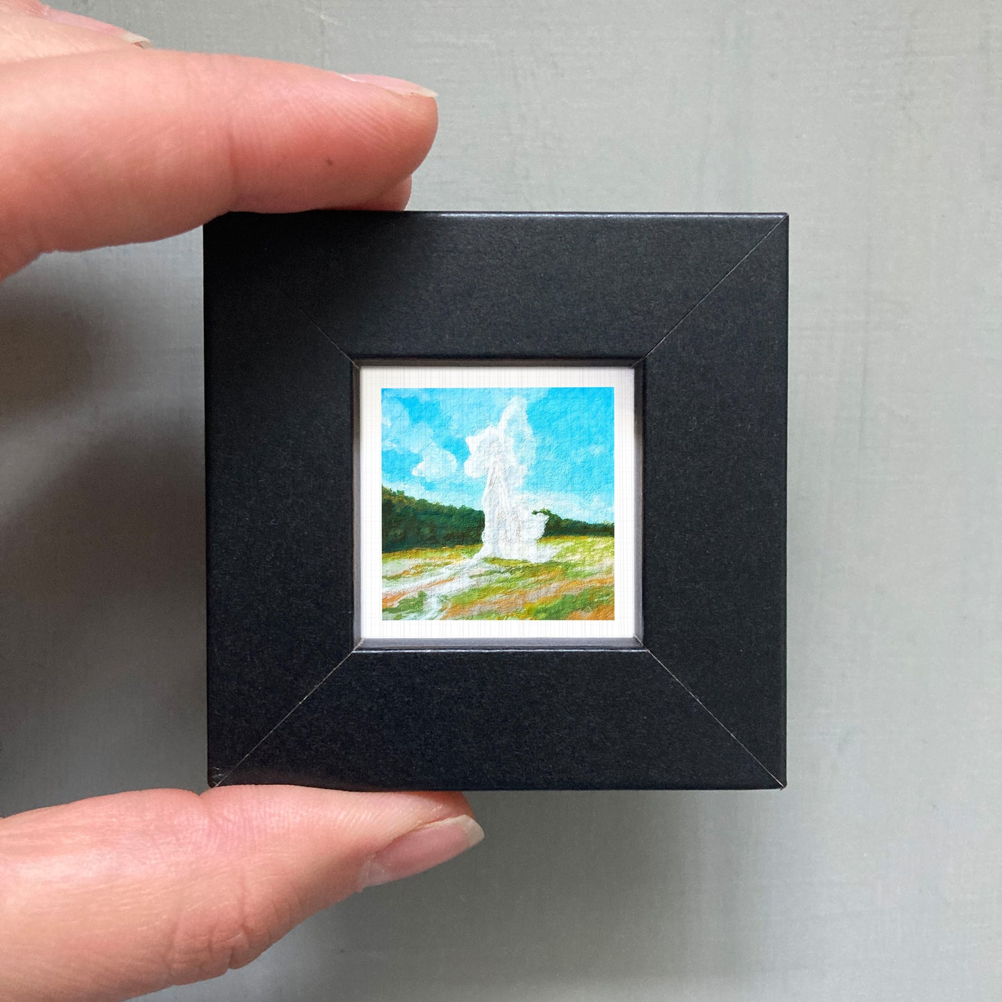 A person's hand holding a small black frame containing a miniature painting of a landscape with a white tower in the background.