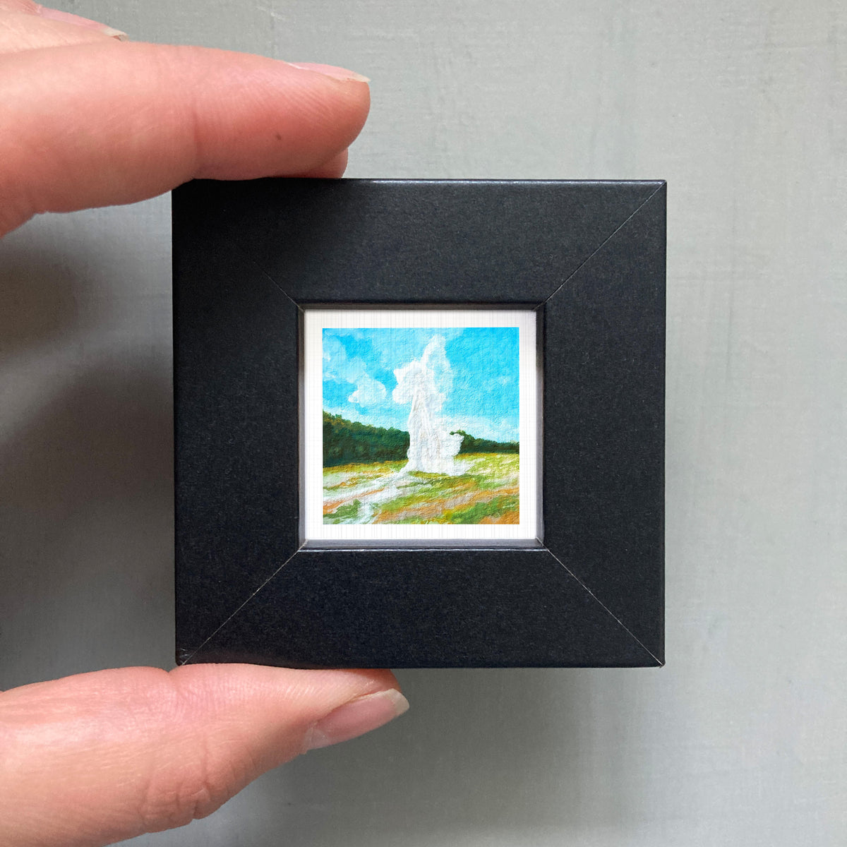 A person's hand holding a small black frame containing a miniature painting of a landscape with a white tower in the background.
