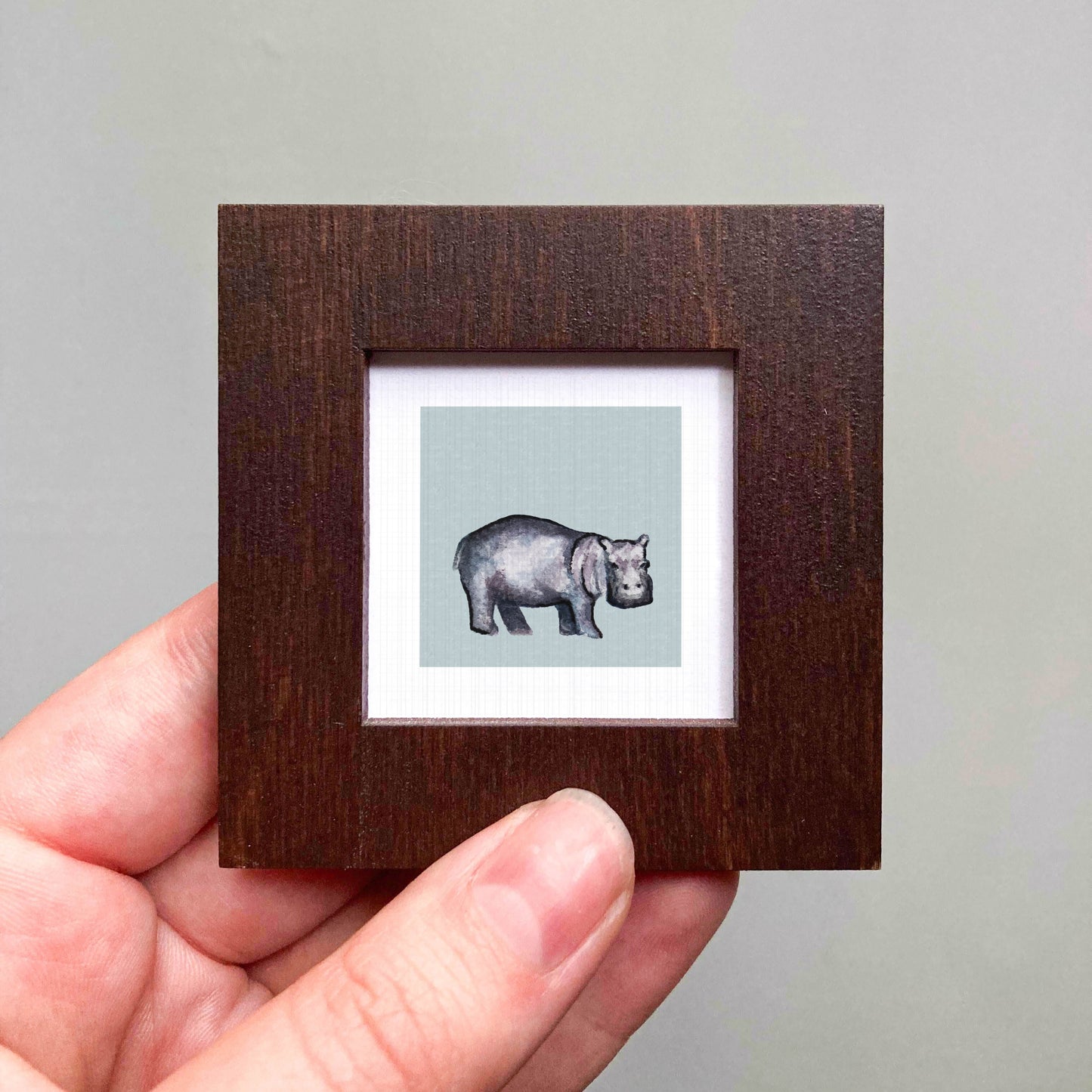 A hand is holding a small wooden frame containing a framed illustration of a hippopotamus.