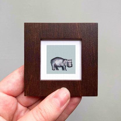 A hand is holding a small wooden frame containing a framed illustration of a hippopotamus.