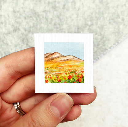 A hand holding a small square picture depicting a field of red flowers against a mountainous background.