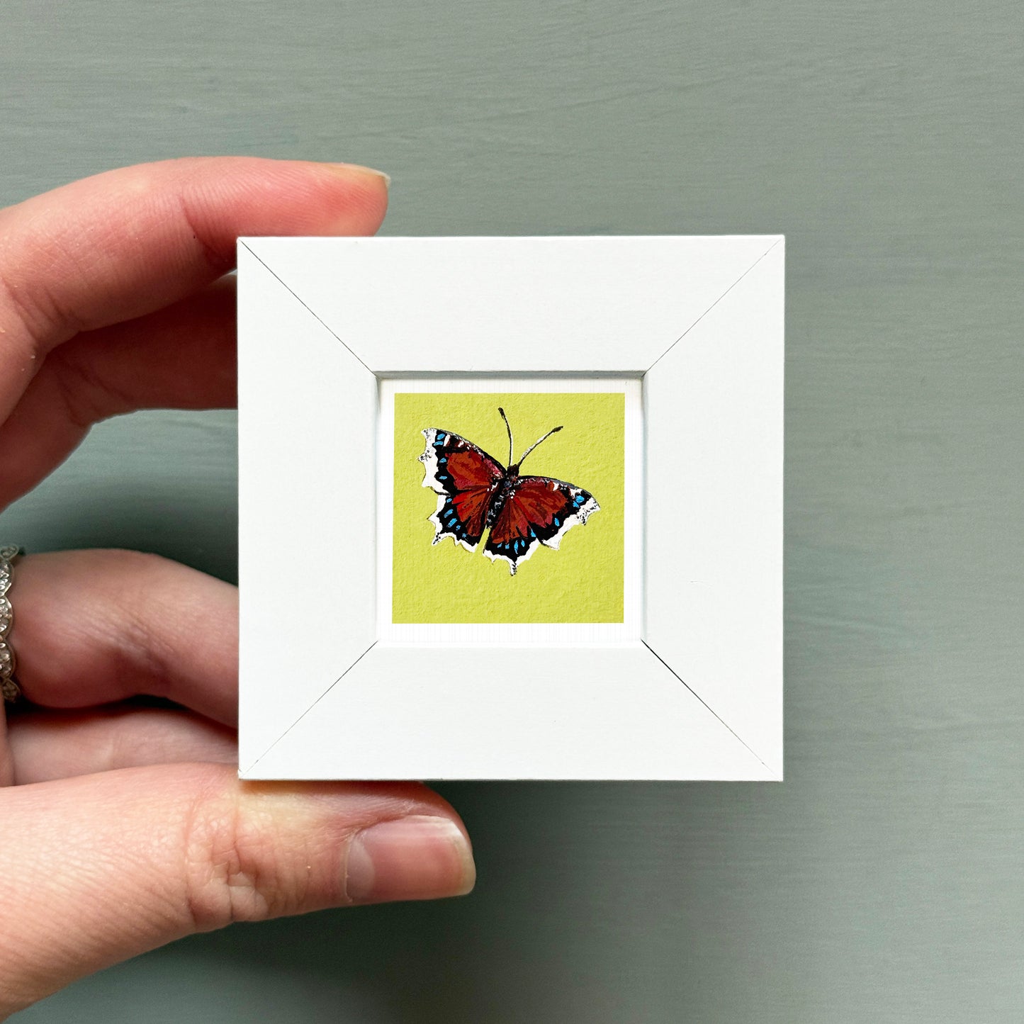 A hand is holding a small white frame containing a colorful painting of a butterfly.