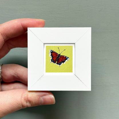 A hand is holding a small white frame containing a colorful painting of a butterfly.