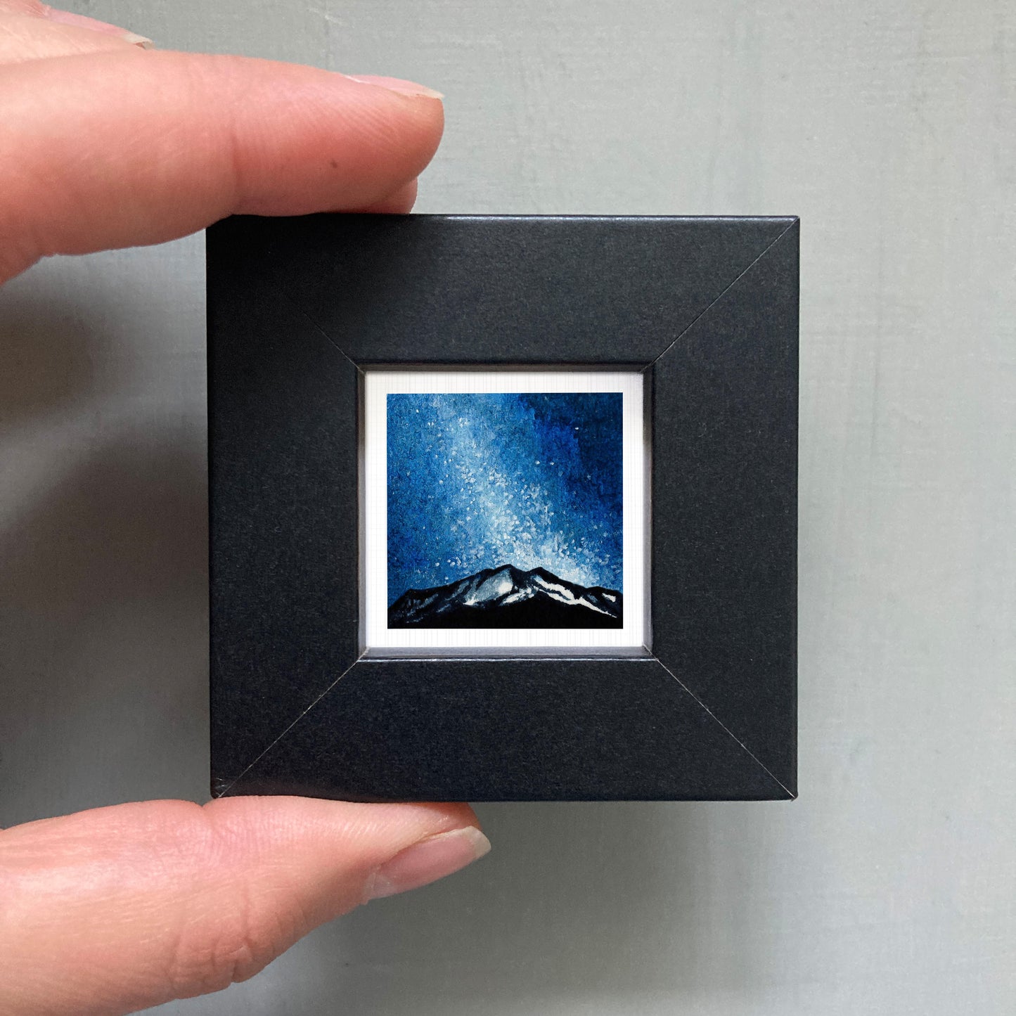 A person's hand holding a small black square frame containing a framed image of a mountain landscape under a starry night sky.