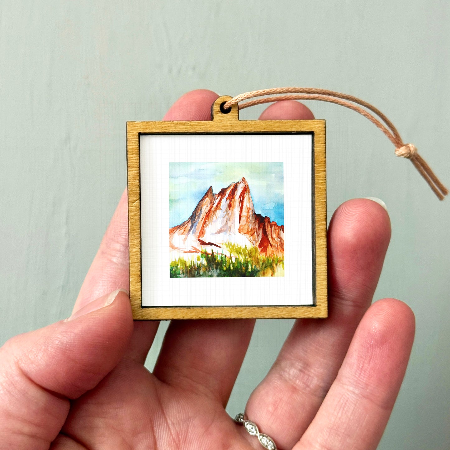A hand holding a small wooden frame containing a watercolor painting of a mountain landscape.