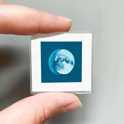 A person's hand is holding a small square with a depiction of a crescent moon on it.