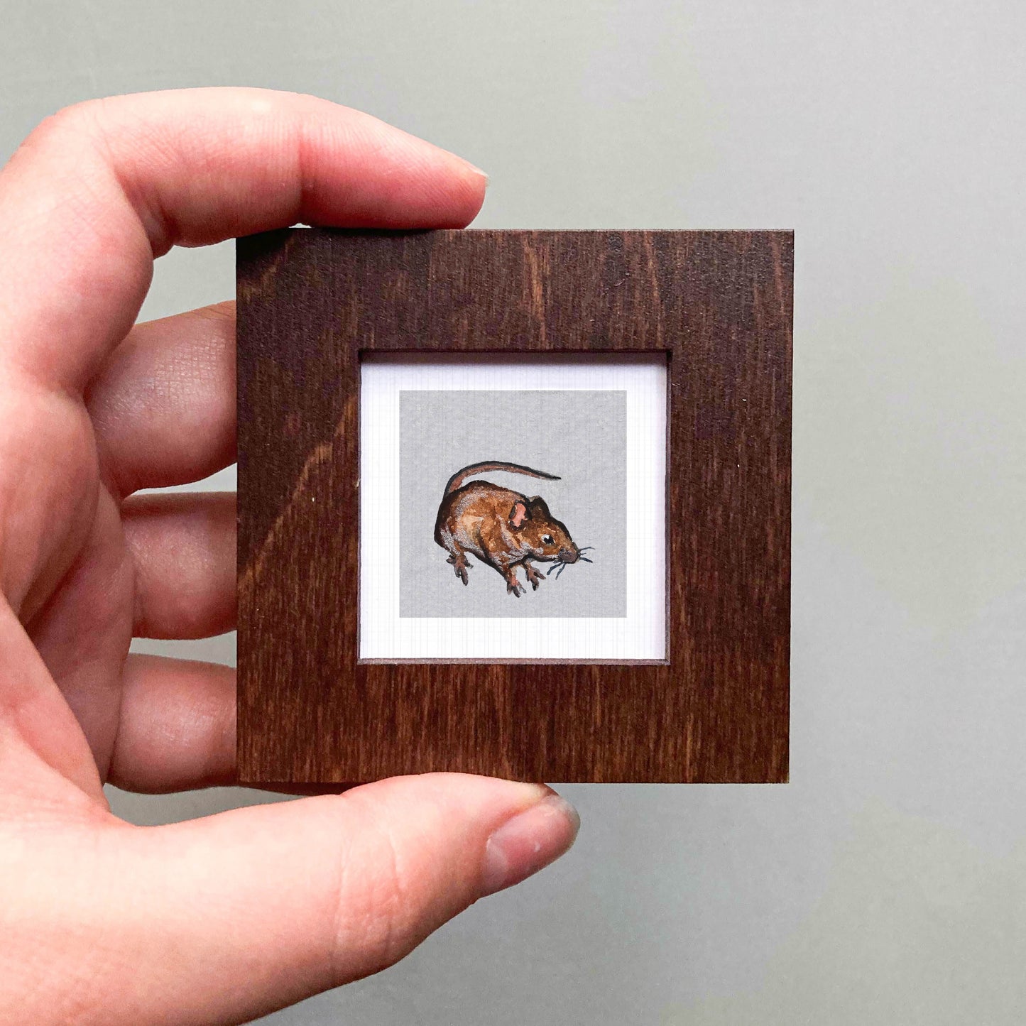 A hand is holding a small wooden frame containing a picture of a brown mouse.