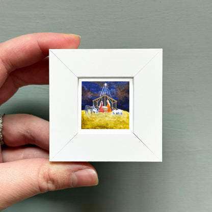 A hand is holding a small white square frame containing a painting of a nativity scene with a man, baby Jesus, and animals in a stable.