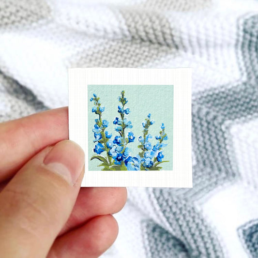 A hand is holding a small square card with a painting of blue flowers on it.