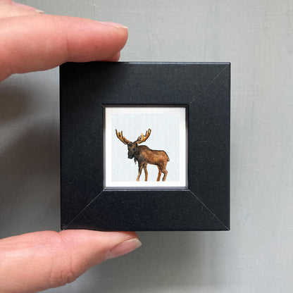 A person's hand is holding a small black picture frame containing a framed image of a moose.
