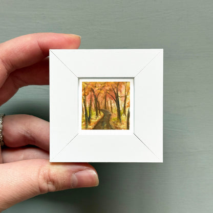 A hand is holding a small white square frame containing a painting of a forest path.