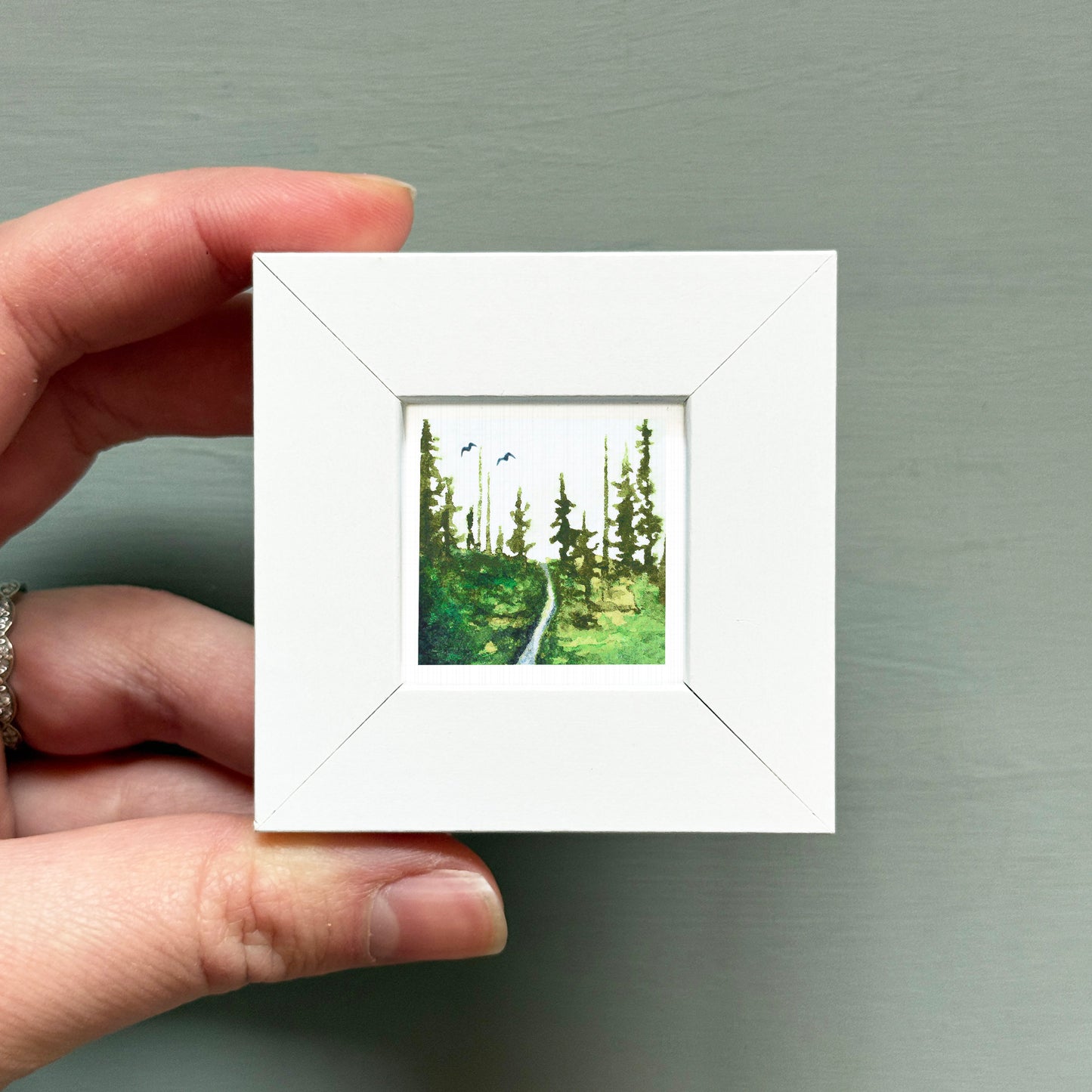 A hand is holding a small white square frame containing a painting of a forest path.
