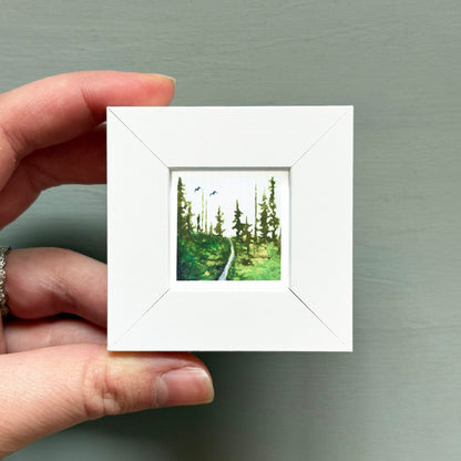 A hand is holding a small white square frame containing a painting of a forest path.