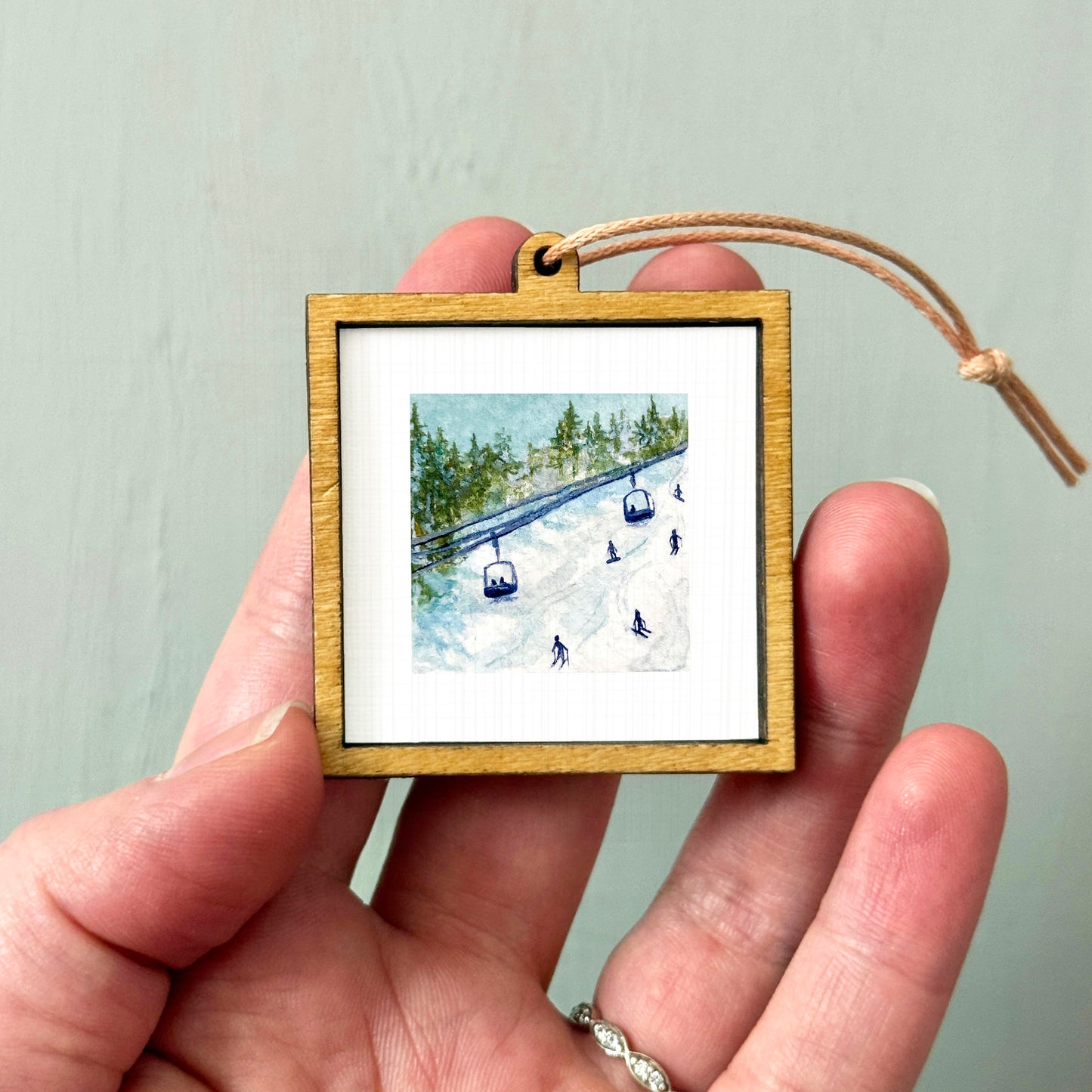 A person's hand holding a small wooden frame containing a framed image of a ski lift and snowy landscape.